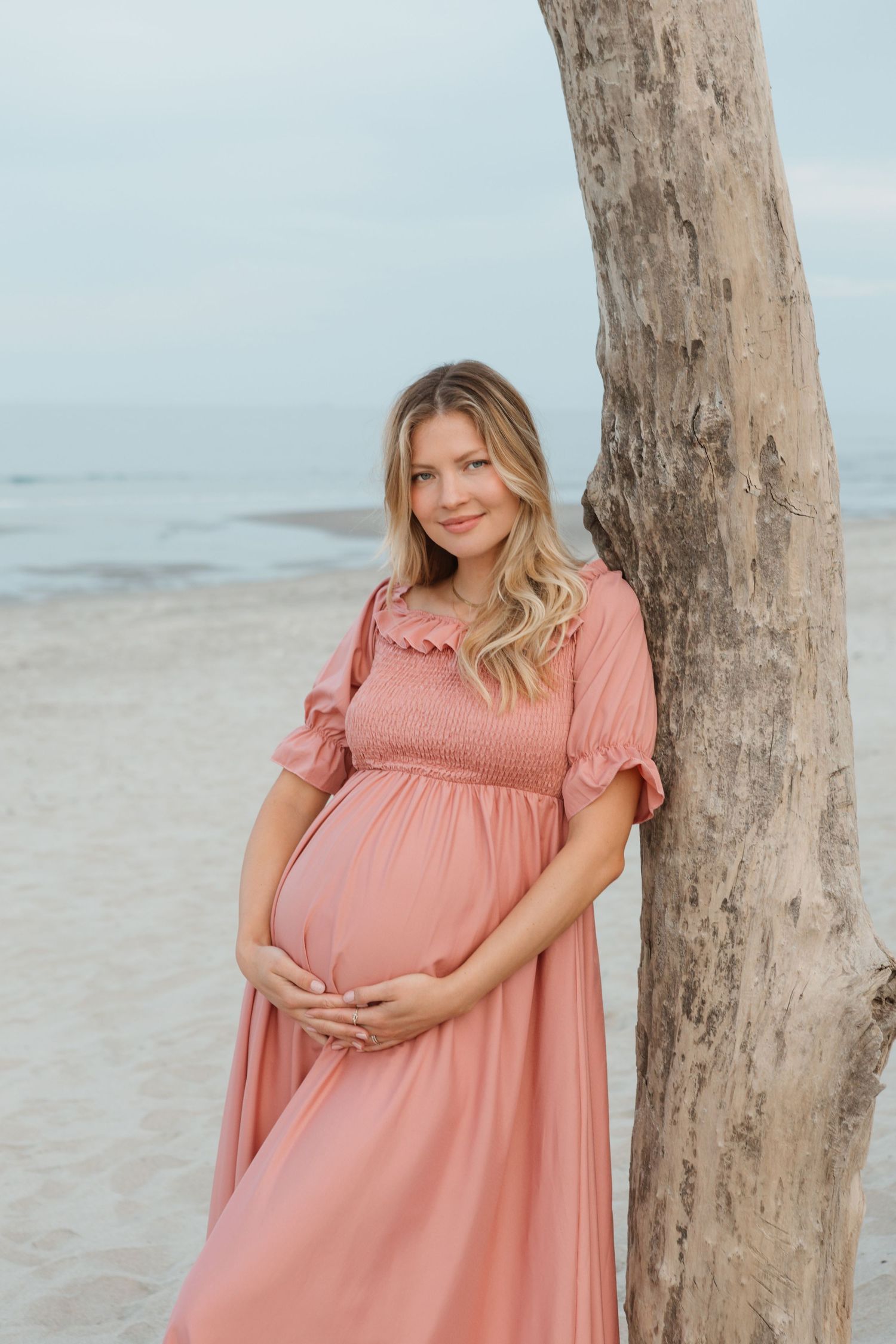 Woman in pink dress cradling baby bump on beach with driftwood tree.