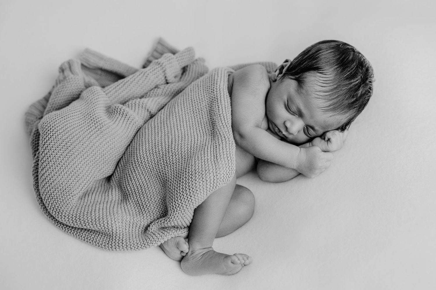 A newborn baby wrapped in a gray knit blanket lying on a white surface in black and white.