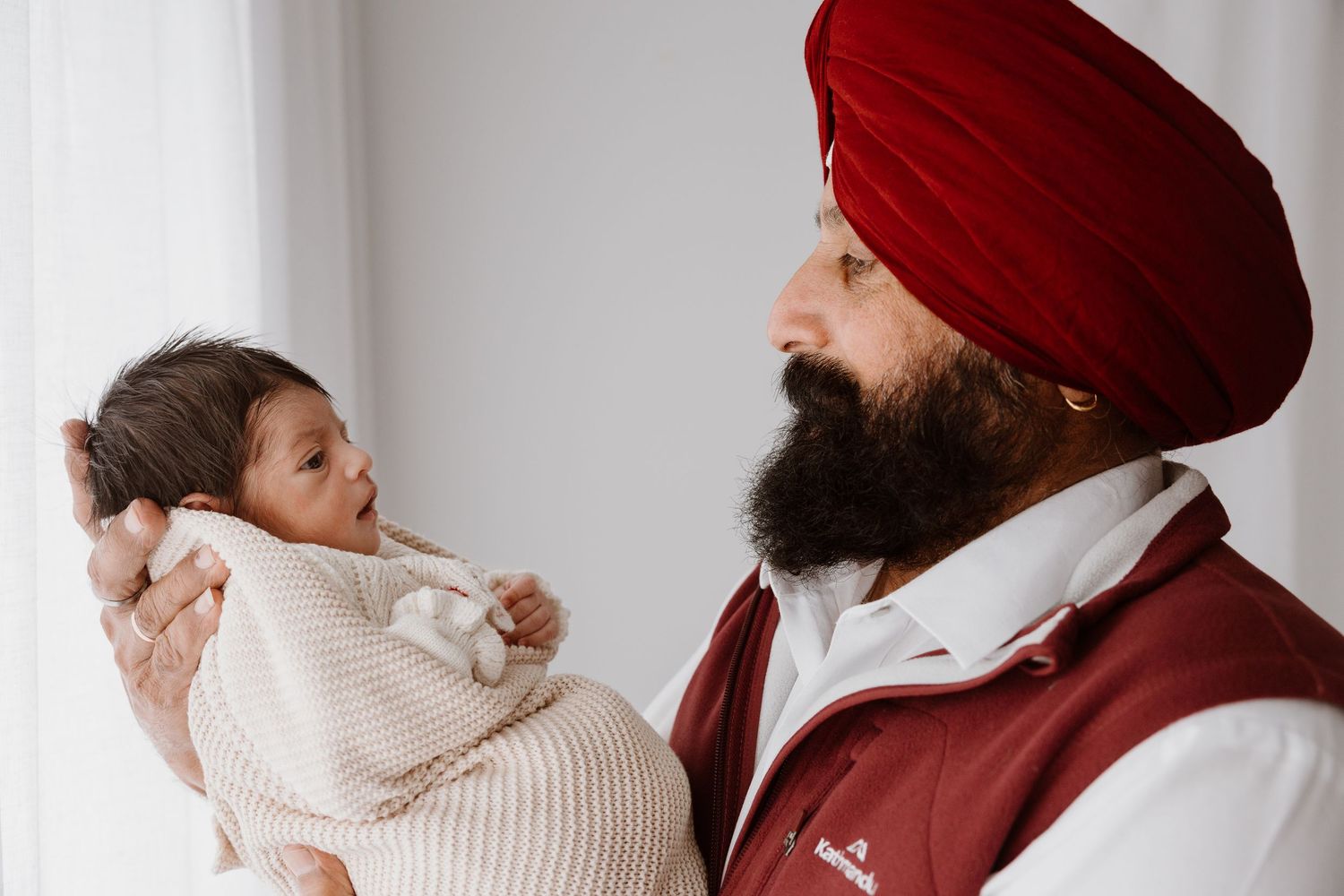 A parent wearing a red turban gently holds a newborn baby wrapped in a white blanket against a light background.