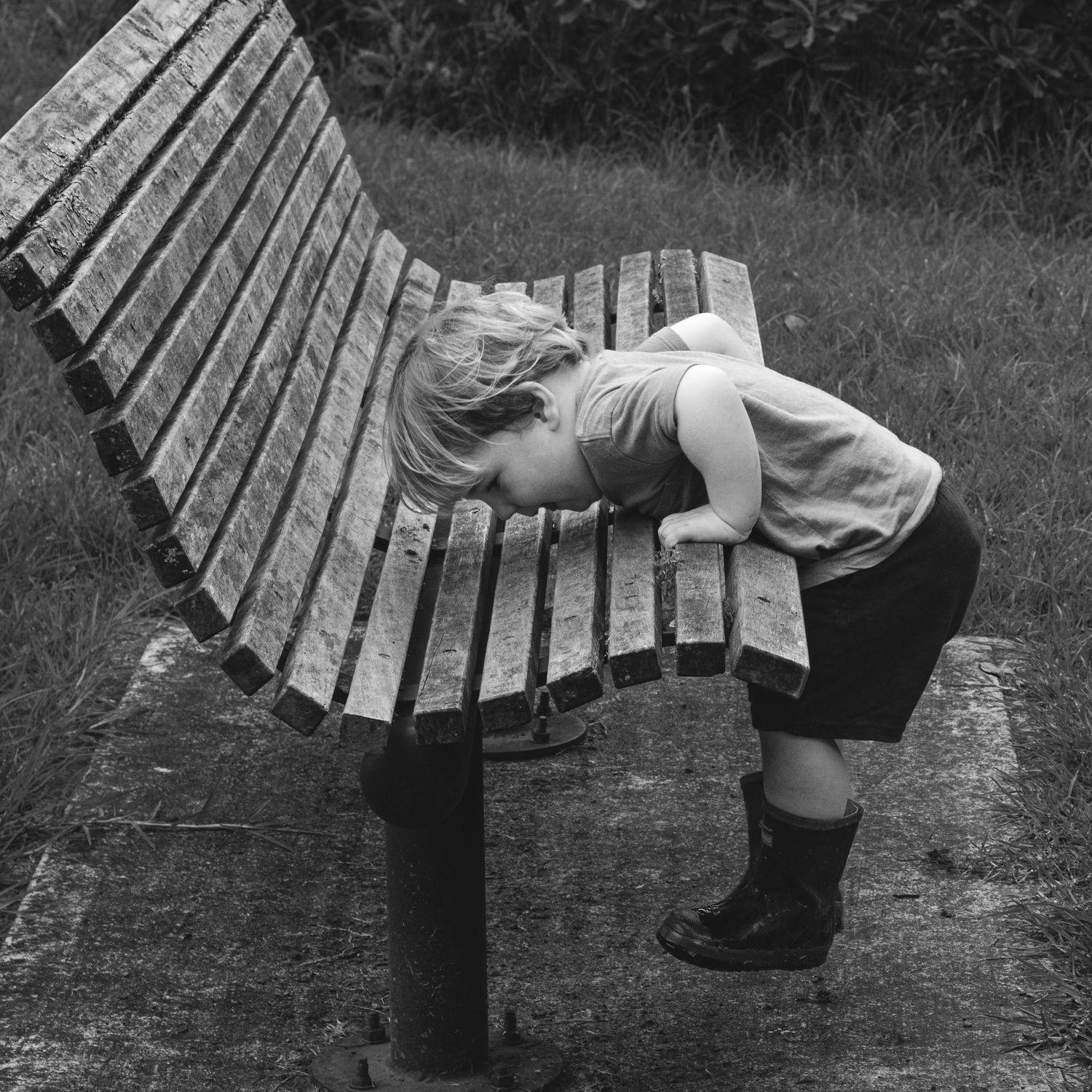 A black and white photo of a small child leaning over a wooden park bench.