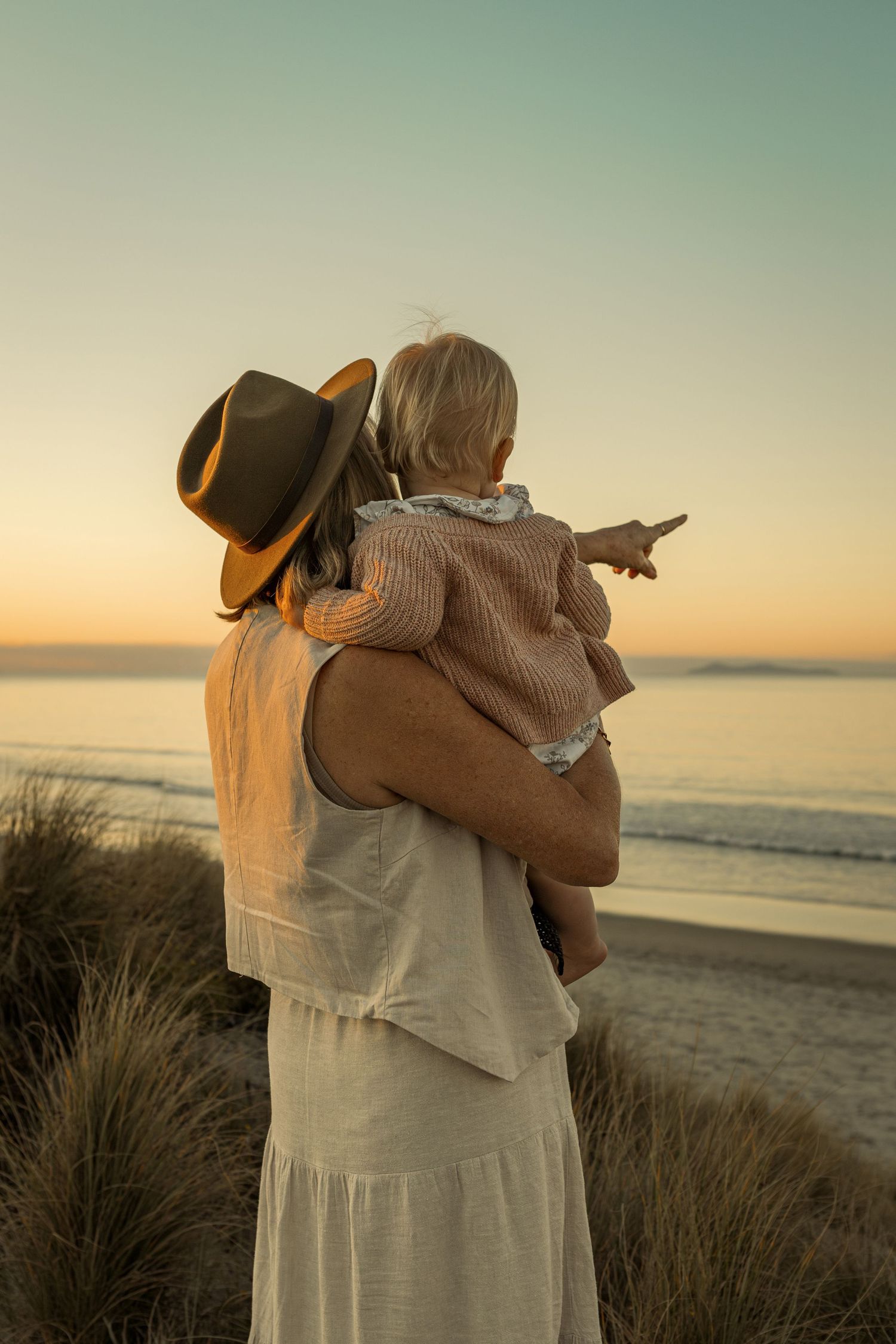 Figure in white dress and hat holding child while pointing at sunset over ocean.