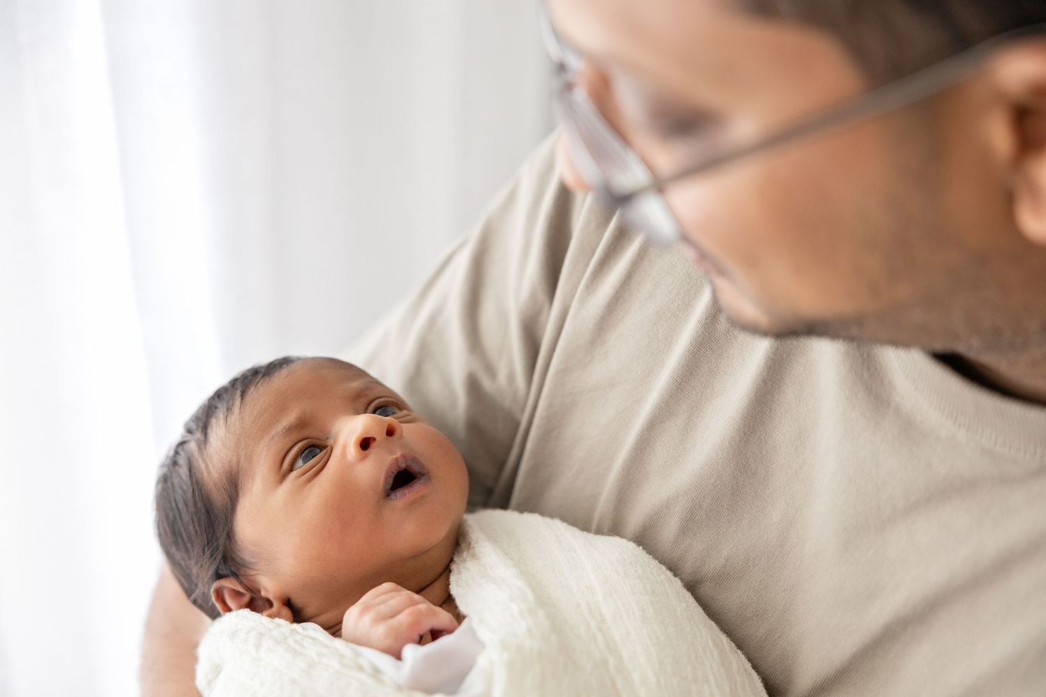 Peaceful moment between a parent and newborn baby wrapped in white blanket, sharing a tender gaze. Newborn photography at home in Tauranga