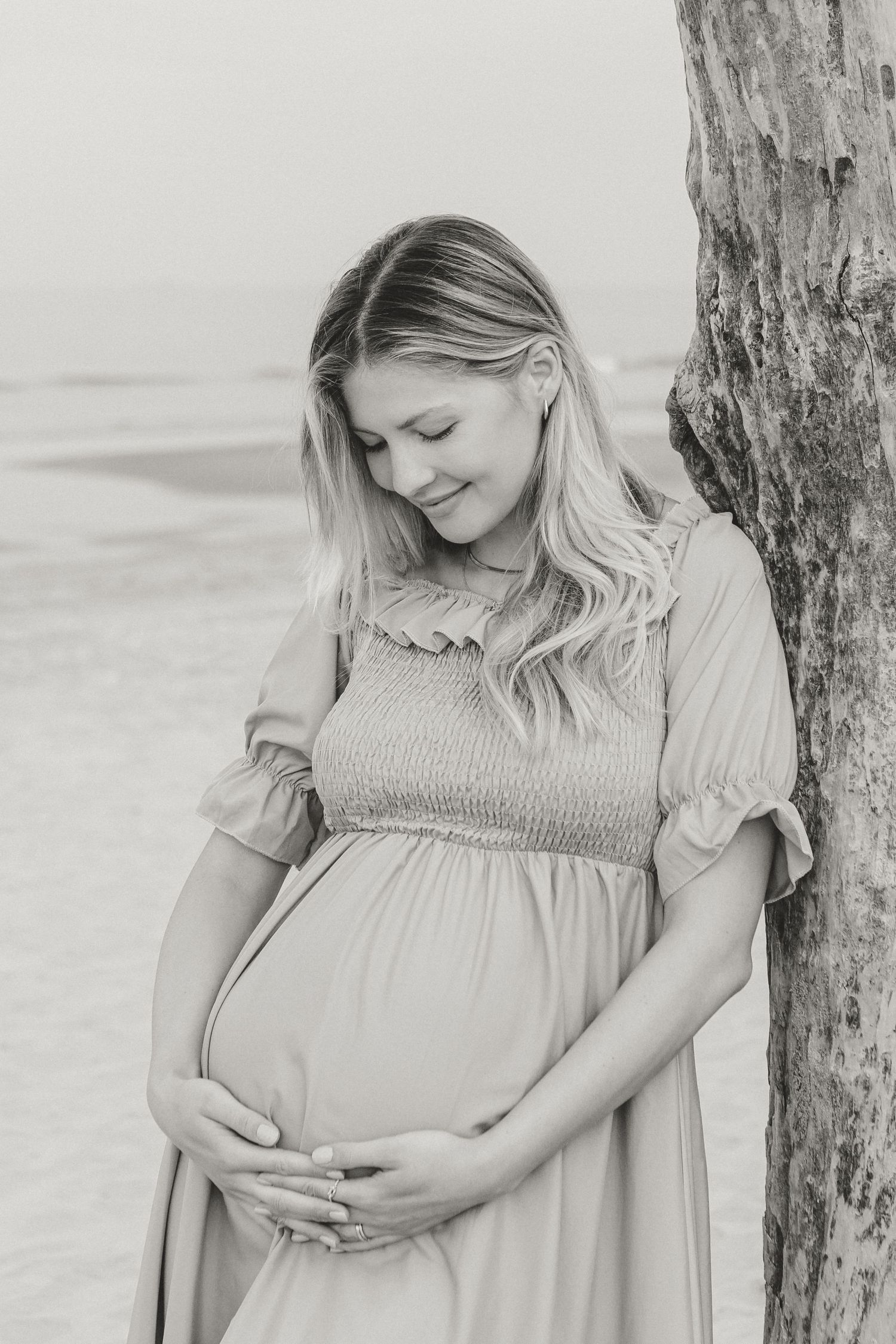 A black and white portrait of an expectant mother leaning against a tree by the ocean.