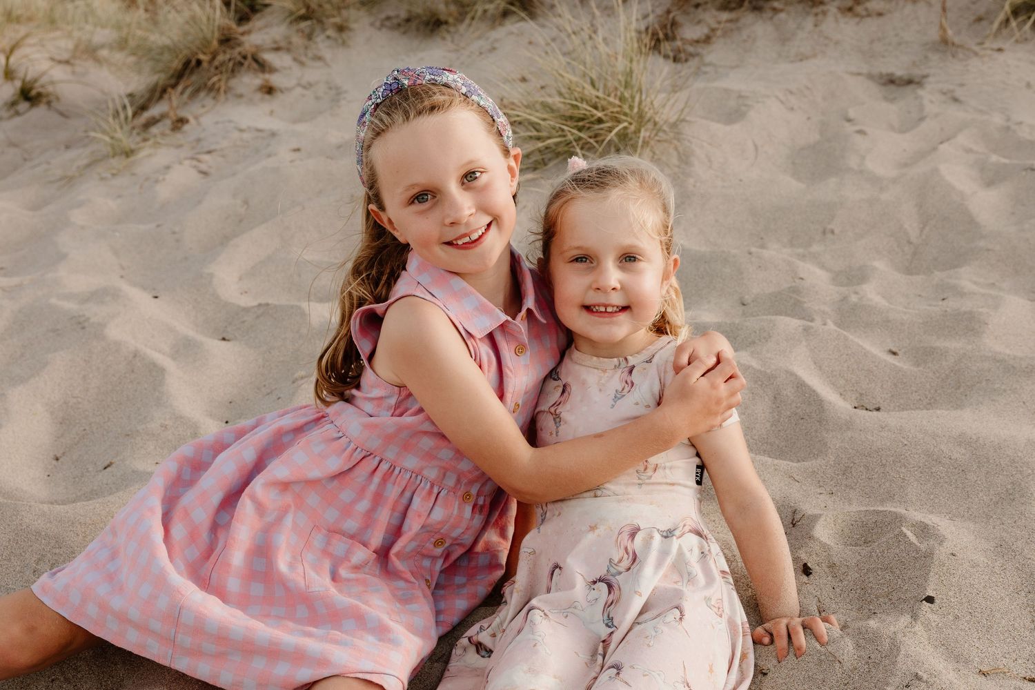 Two young children in floral dresses sitting together on sandy beach during sunset.
