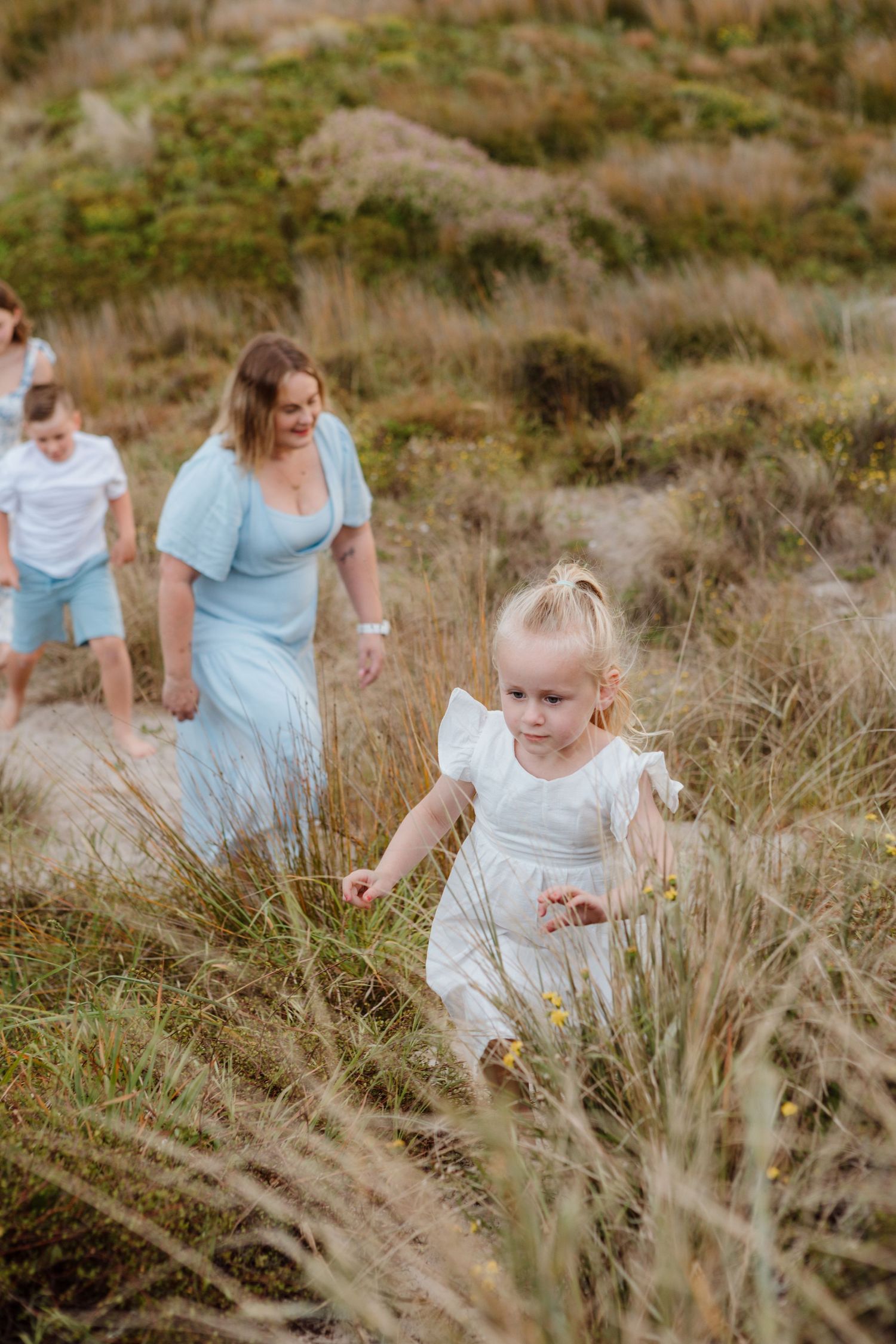 A white-dressed figure explores a grassy coastal trail as others follow behind in soft evening light.