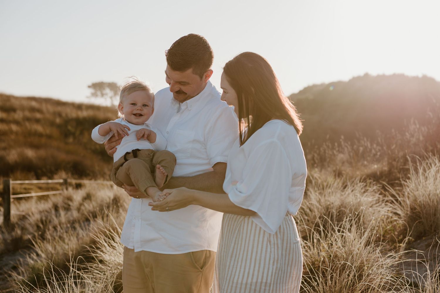 Young couple at sunset holds their baby while standing in golden grass field during warm summer evening. Tauranga photographer Papamoa Beach