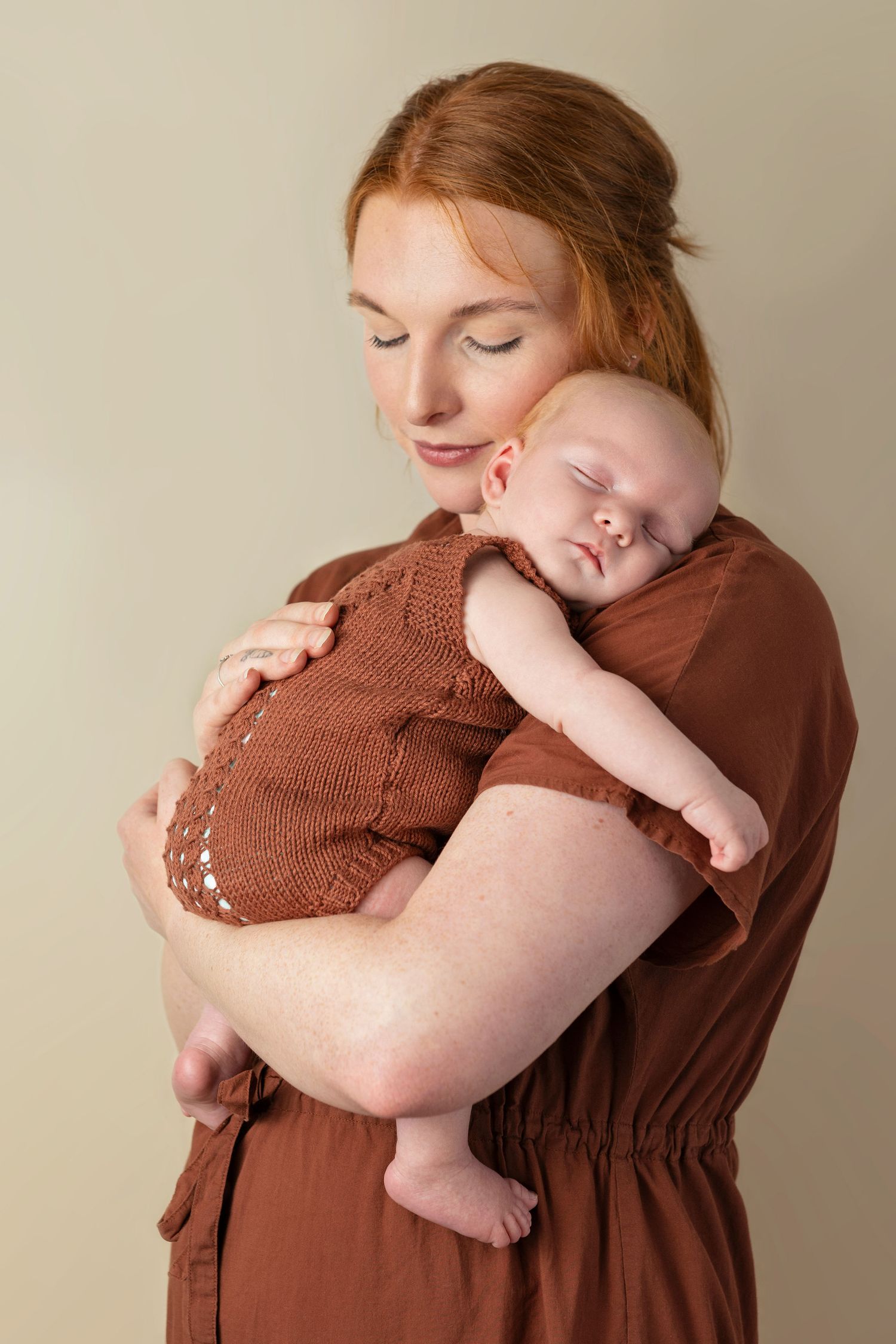 A tender moment captured as a sleeping baby in a brown knit romper rests peacefully against a parent in earth tones.