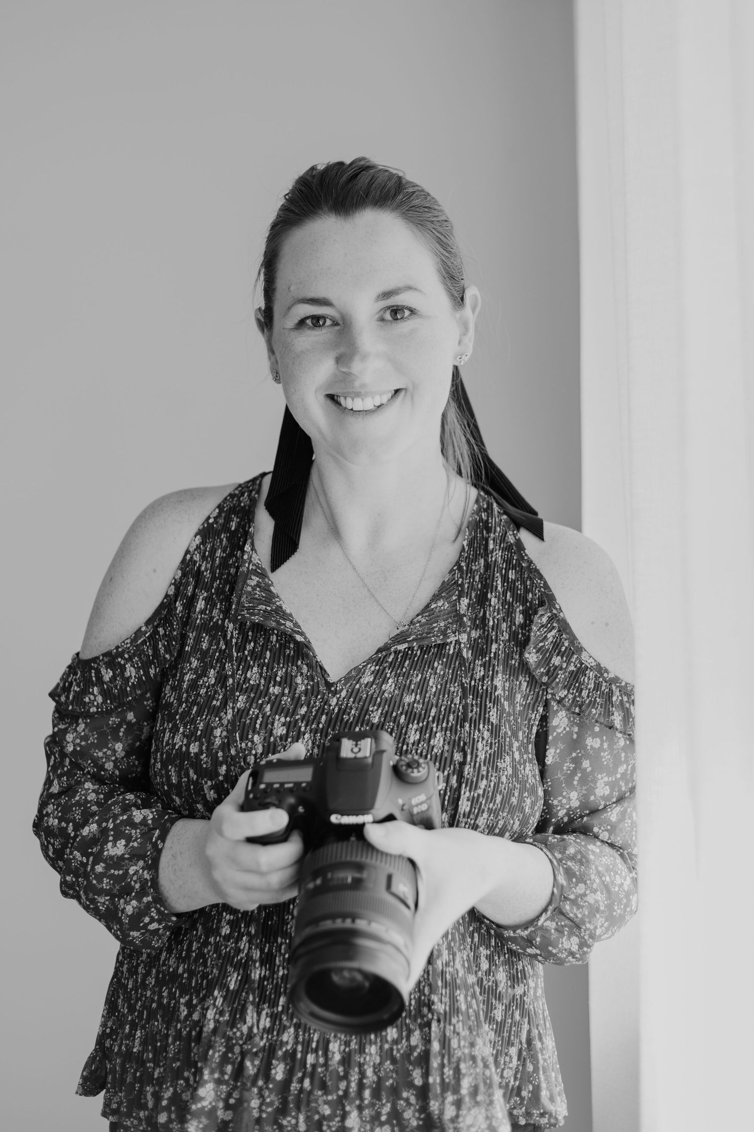 Black and white portrait of a photographer in a patterned dress holding a professional camera against a light background. Tauranga headshot