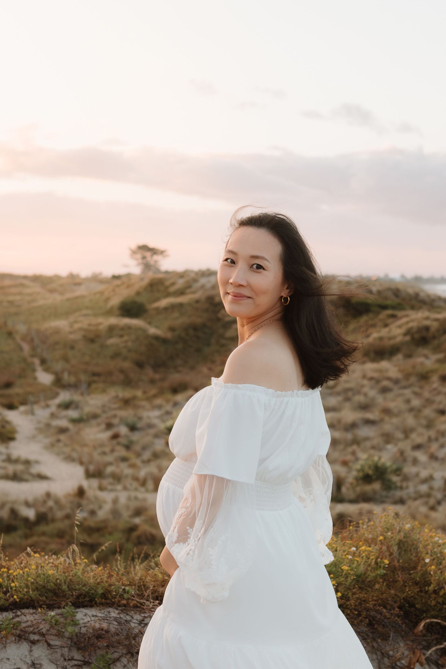 Woman in a flowing white dress poses on a hill during golden hour.
