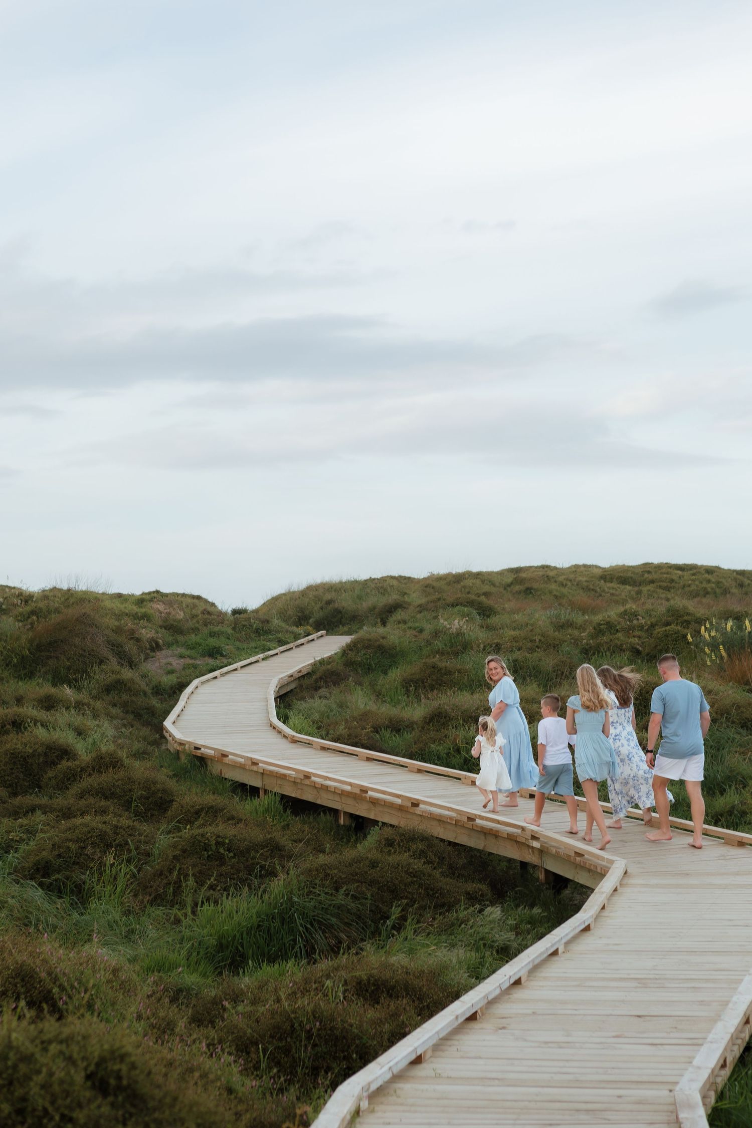 A family walks along a winding wooden boardwalk through coastal dunes under cloudy skies.