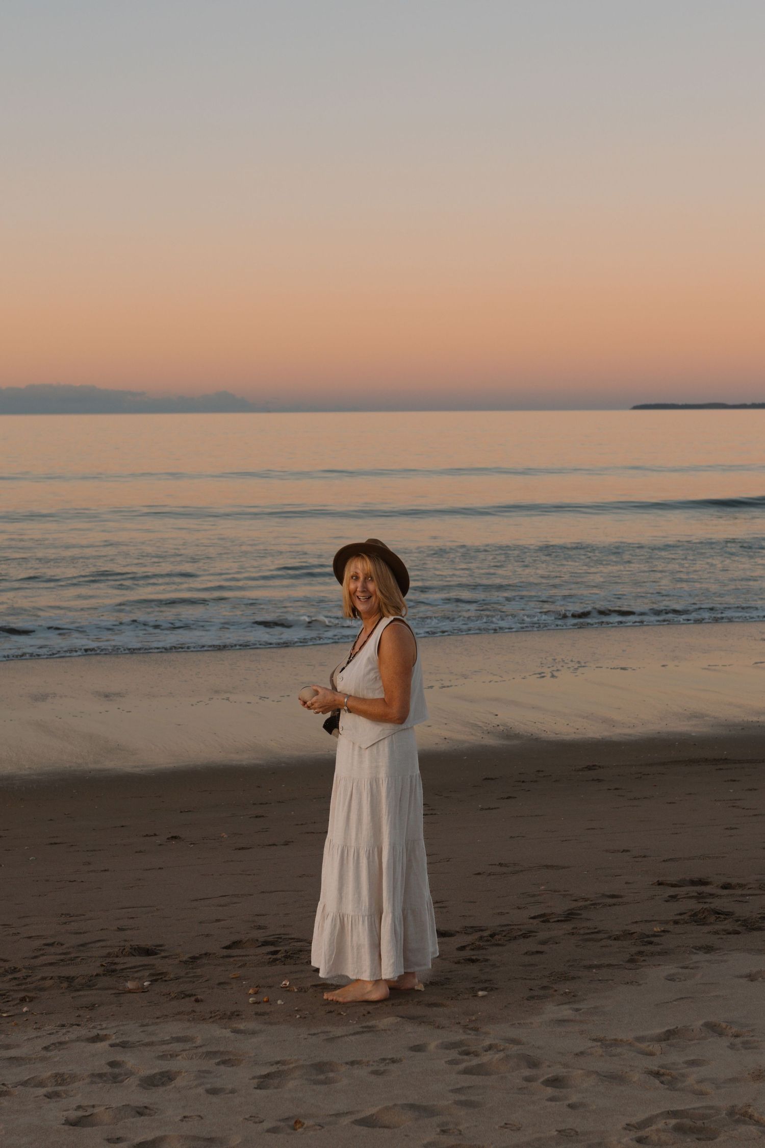 Woman in white dress and hat stands on empty beach during golden hour sunset.