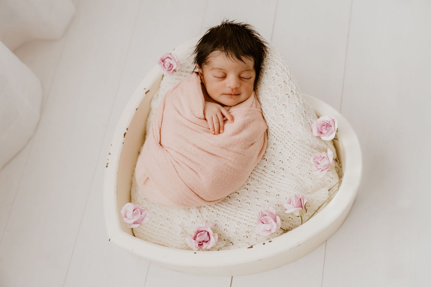 A sleeping newborn wrapped in pink, nestled in a white bowl decorated with pink roses.