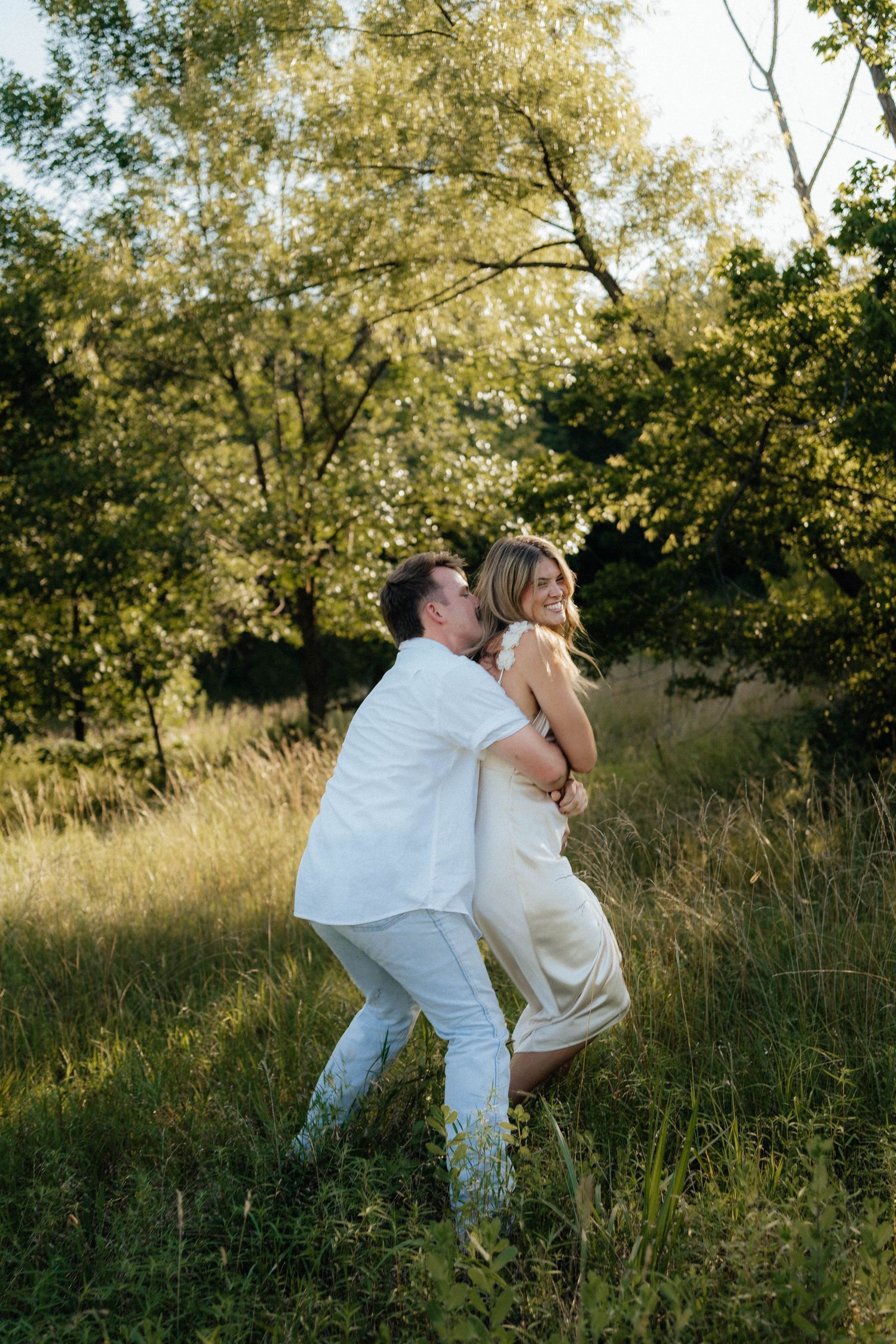 A couple in white outfits dances together in a sun-dappled meadow during golden hour, creating a romantic moment.