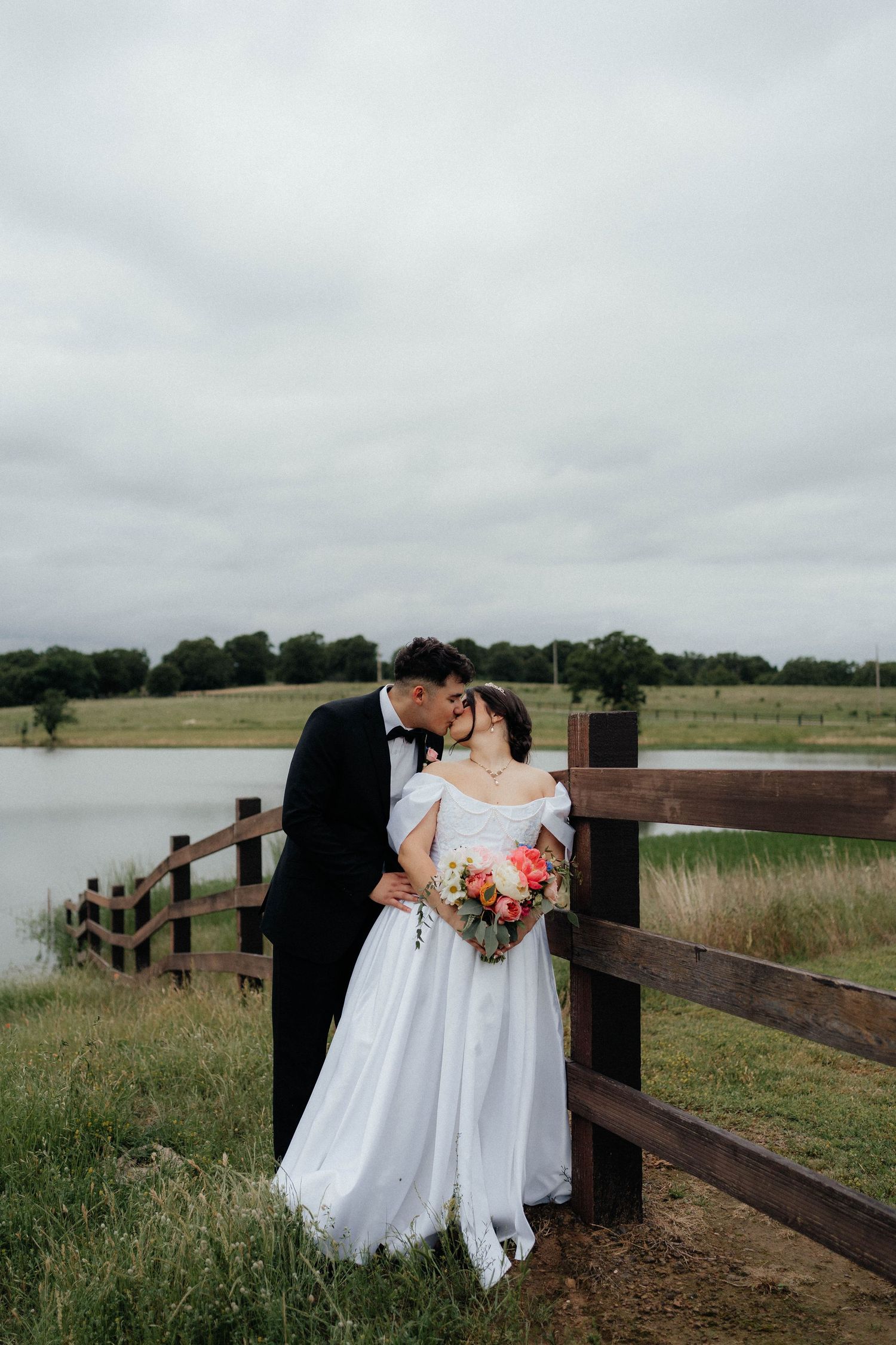 A romantic wedding couple embraces by a rustic wooden fence overlooking a serene pond on an overcast day.