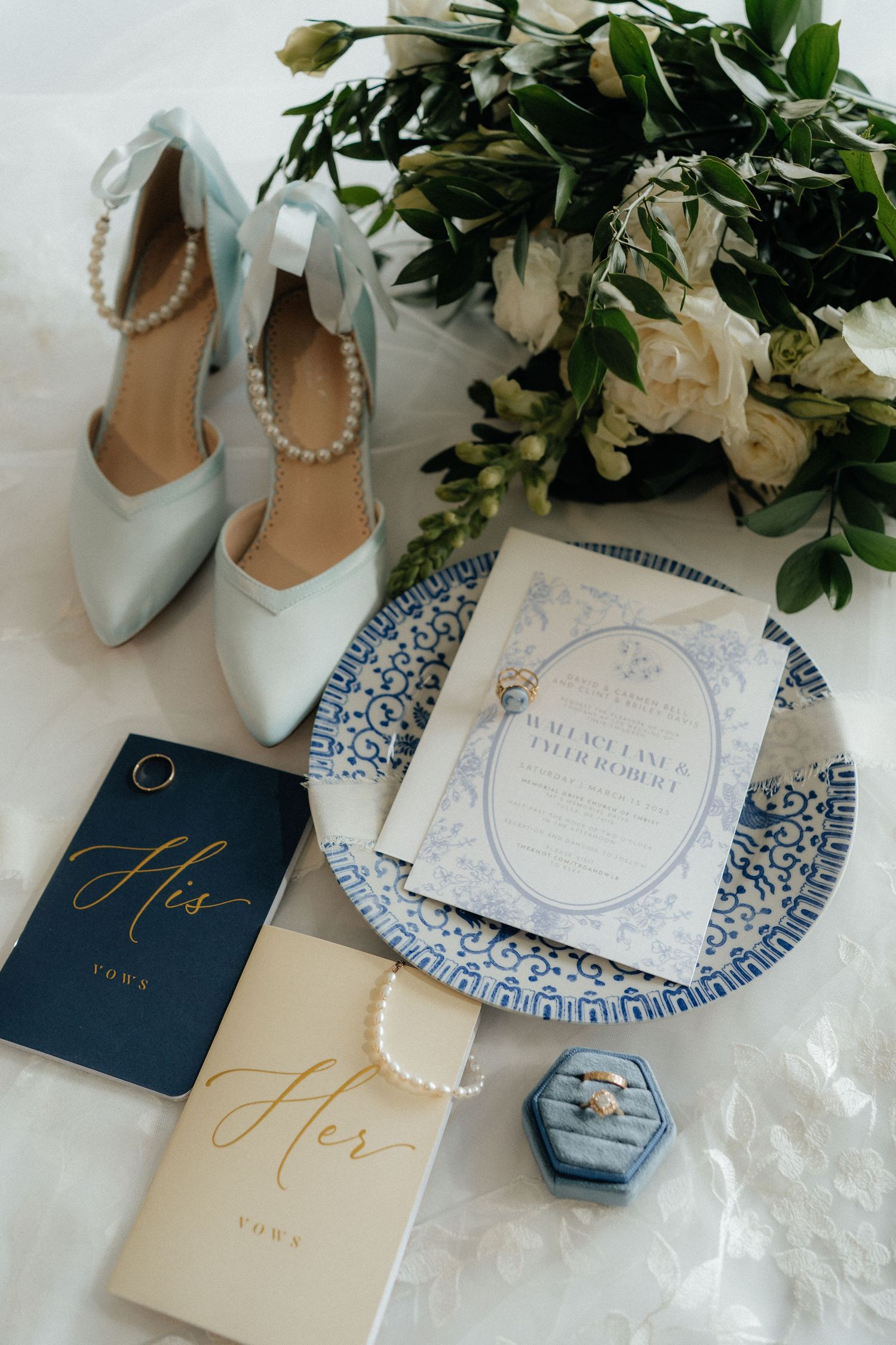 Wedding details flatlay with blue stationery, pearl-adorned shoes, and white floral arrangement.