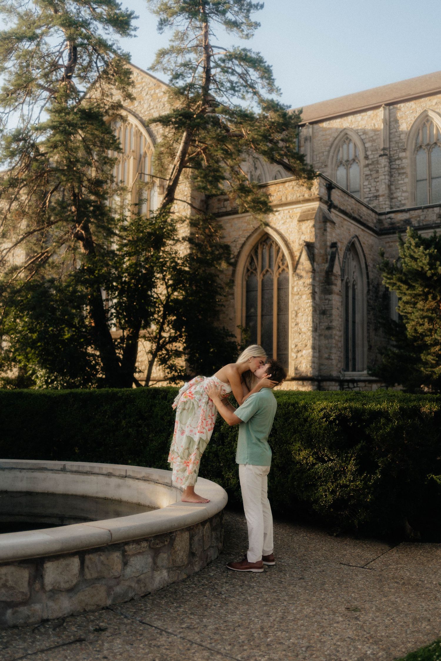 A couple shares an intimate moment by a stone wall outside a Gothic-style church with arched windows and tall pine trees.