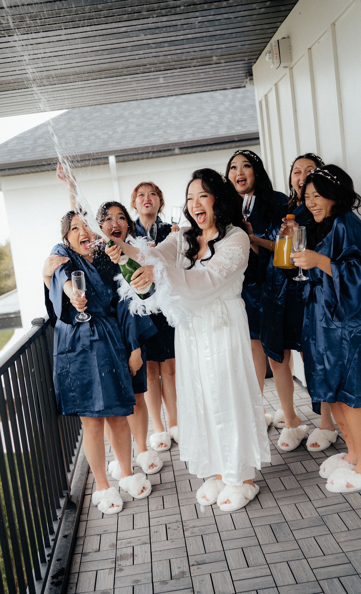 Bridal party in blue robes celebrates with champagne and laughter on a balcony.