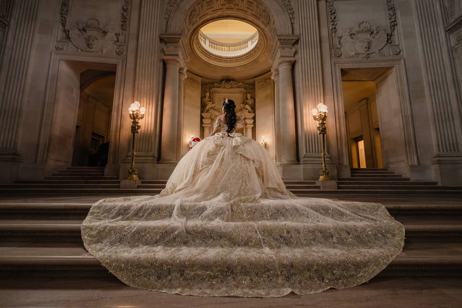 A luxurious quinceanera dress with a dramatic train sprawls across stone steps in an ornate classical building with warm lighting.