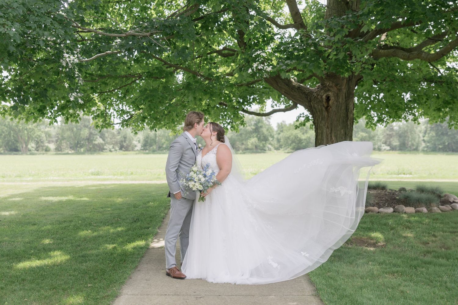 Wedding couple in a beautiful garden setting poses while veil flows gracefully in the breeze.