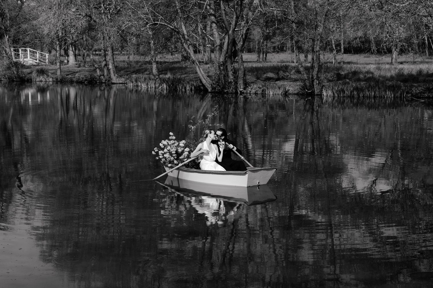 A romantic black and white wedding photo series shows newlyweds in a boat on a tranquil lake surrounded by trees.