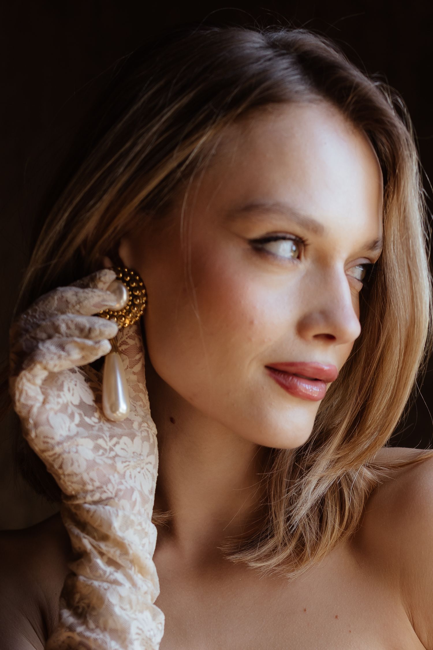 A softly lit portrait highlighting delicate lace gloves, vintage jewelry, and gentle side glance expression.