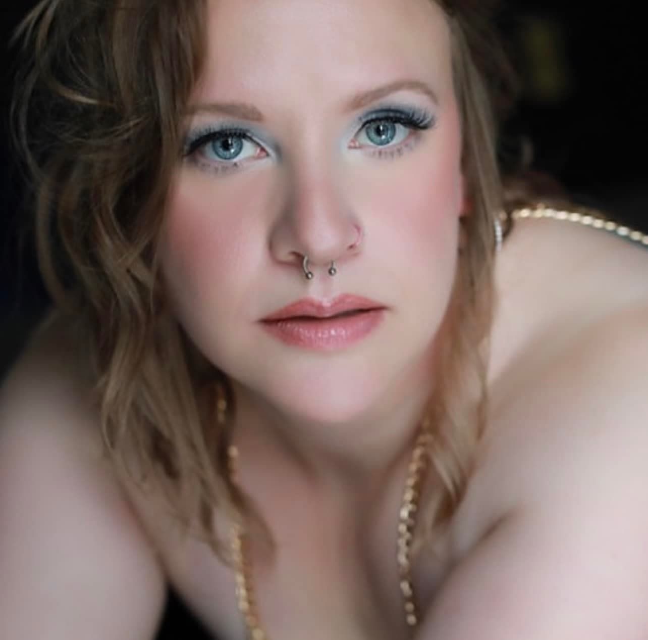 Portrait photo showing striking blue eyes, a septum piercing, and soft pearl necklace in moody lighting.