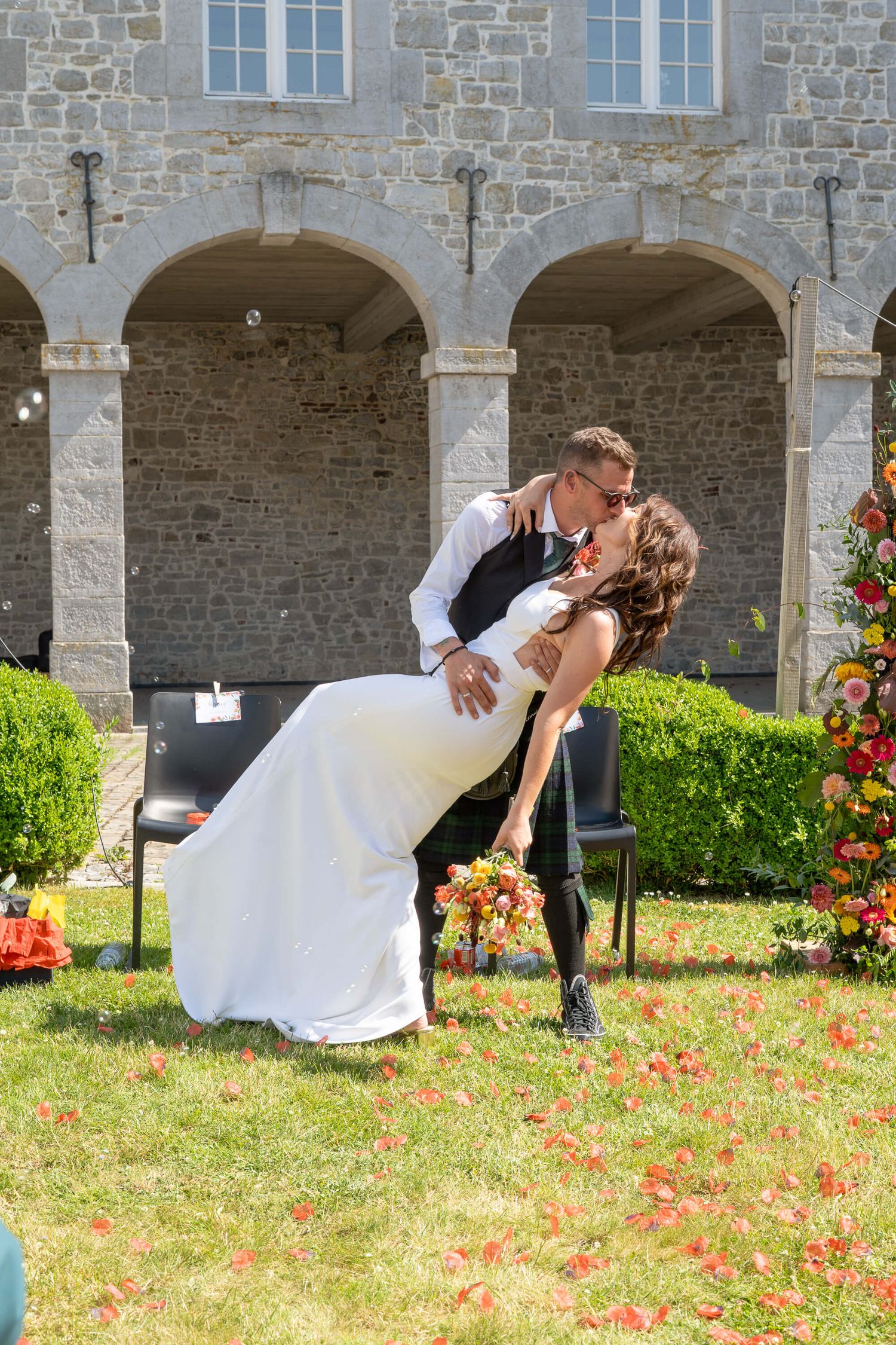 Romantic wedding couple shares a passionate dip kiss in front of stone archways and colorful garden on sunny day.