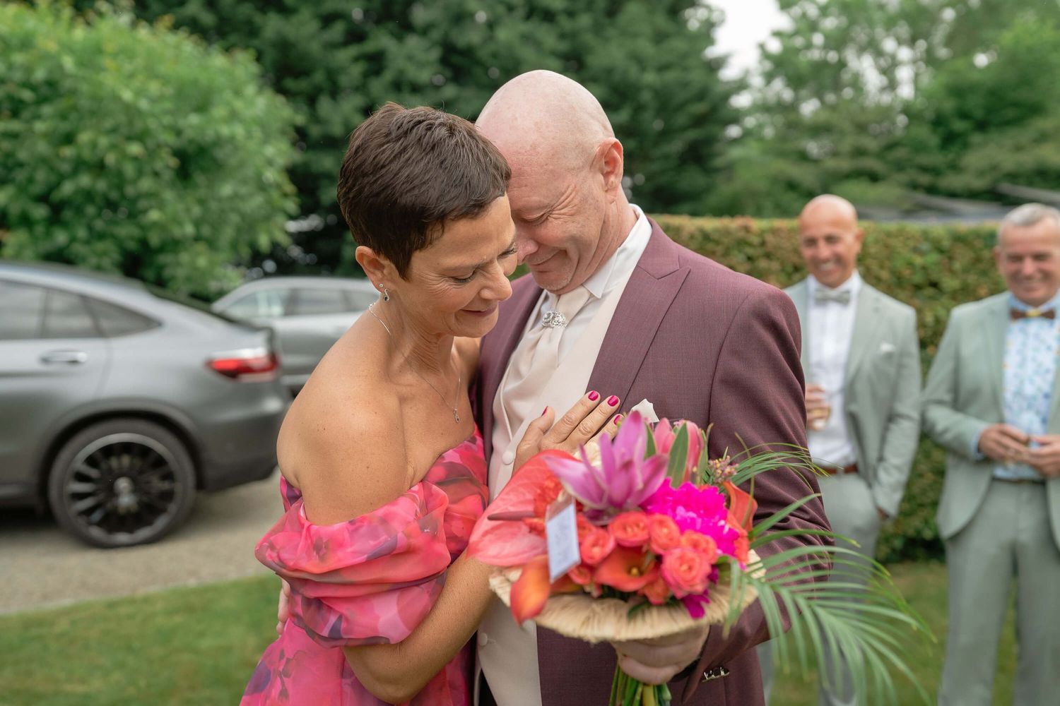 Couple shares intimate moment at outdoor wedding ceremony while holding vibrant pink and tropical floral bouquets.
