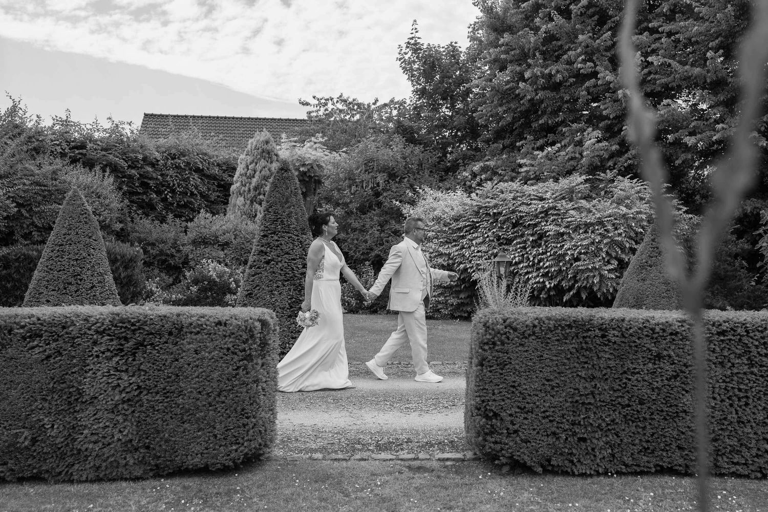 A couple in wedding attire holds hands while walking between neatly trimmed garden hedges in black and white.