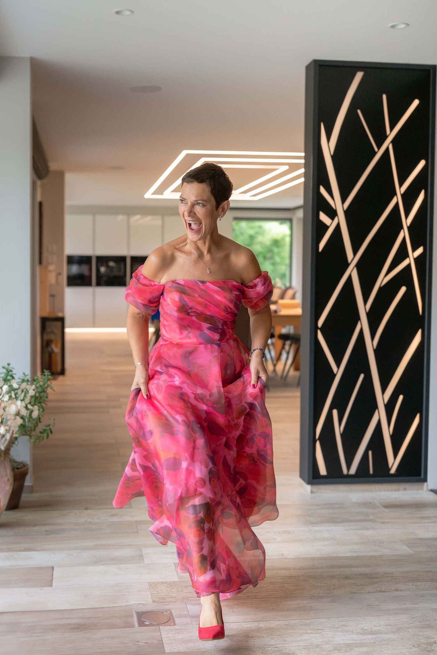 A flowing pink floral off-shoulder gown is modeled in a modern interior space with geometric lighting and wood floors.