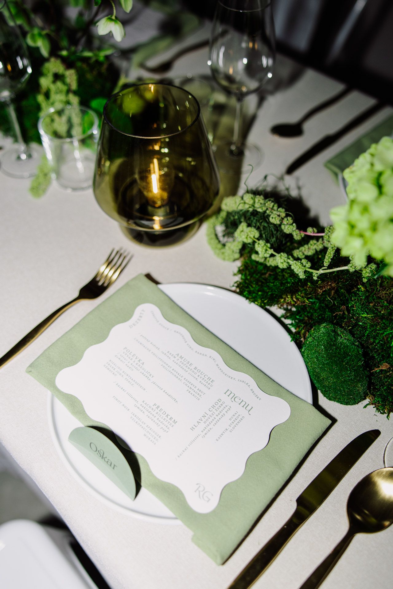 An elegant table setting with a sage green menu card on white plate, wine glass and fresh greenery decorations.