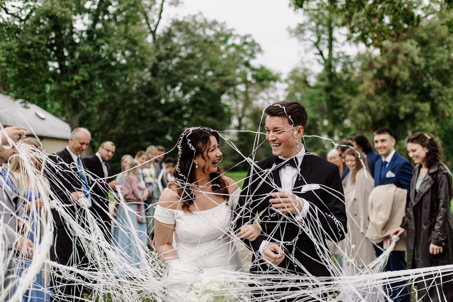 Couple laughs and celebrates while guests shower them with white streamers during outdoor wedding ceremony.