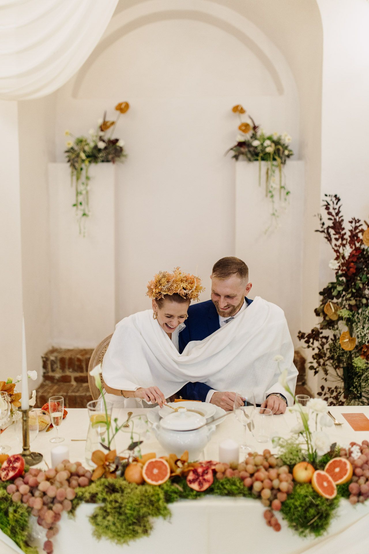 Couple wrapped in white blanket dining at rustic table decorated with citrus and greenery.