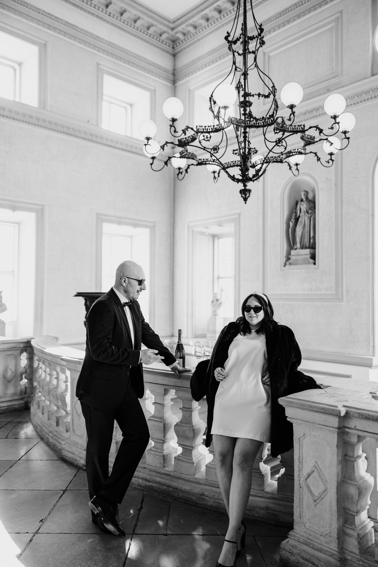 Black and white photo of two people in formal attire standing by an elegant balustrade under an ornate chandelier.