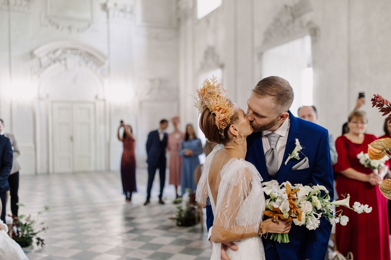 Romantic wedding moment in elegant white baroque hall with guests watching.