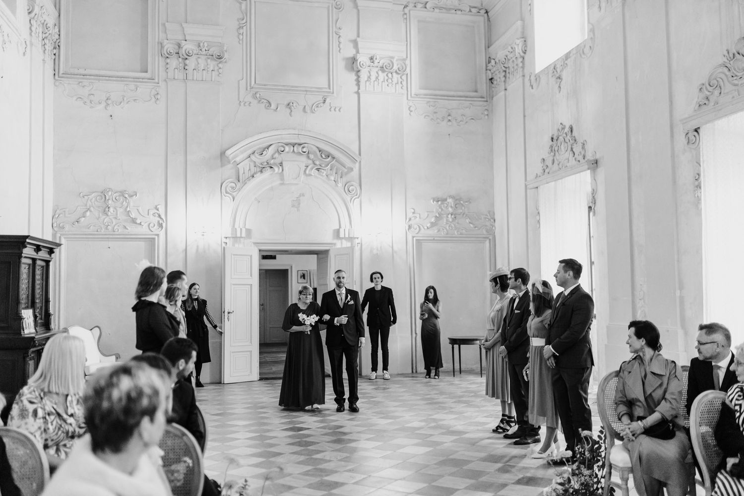 Elegant wedding ceremony in ornate white baroque hall with guests gathered.