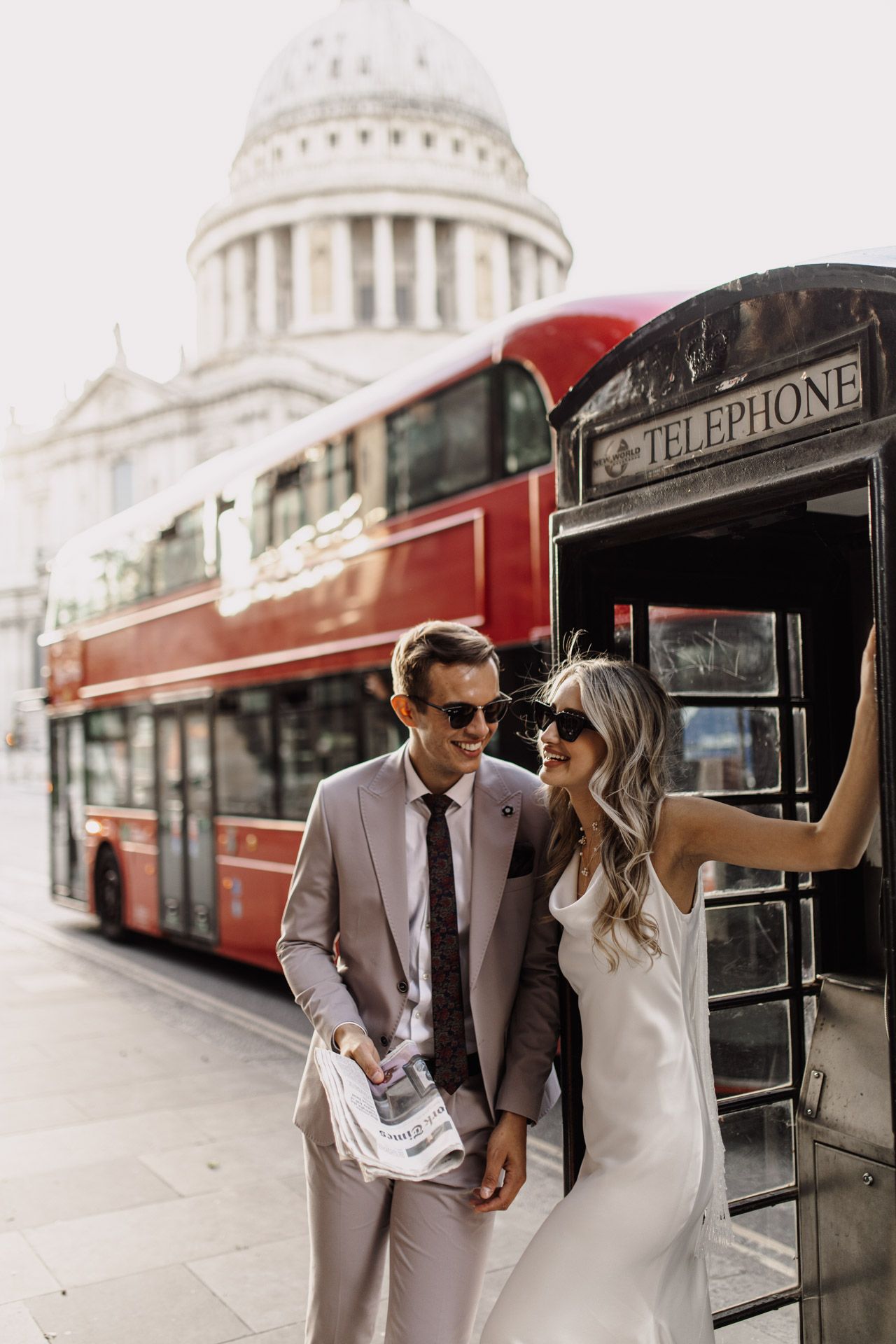 Stylish couple poses by vintage London phone booth with red double-decker bus and St Paul's Cathedral in background.