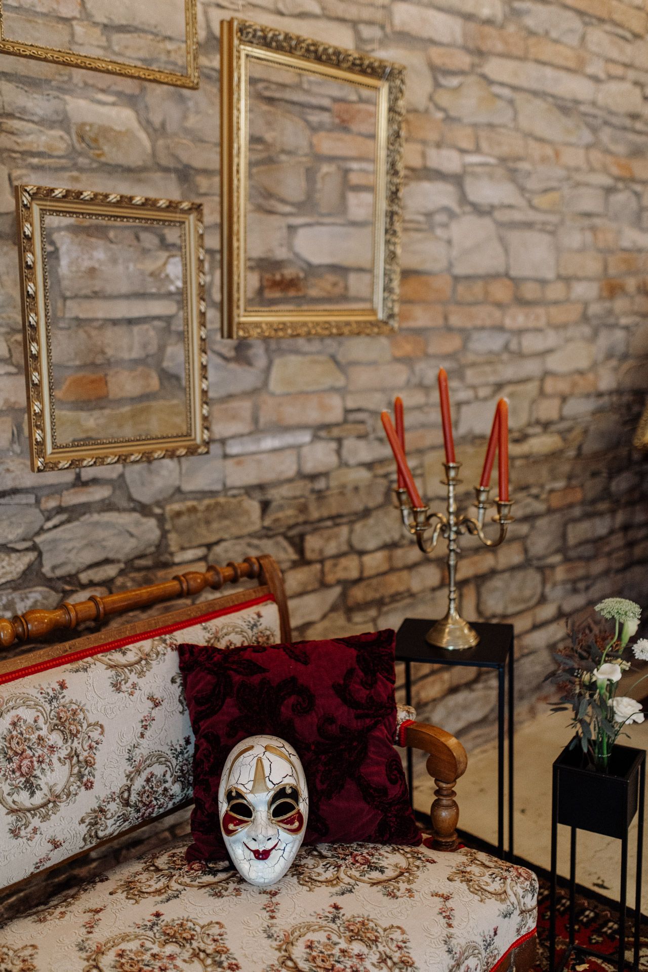 Vintage room decor with ornate gold frames, red candles, and decorative mask on patterned chair.