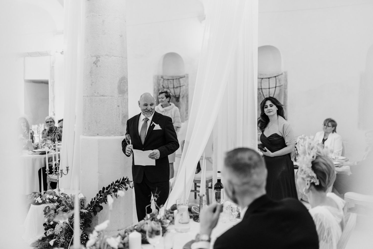 Black and white photo of wedding reception speech with guests seated at decorated tables.