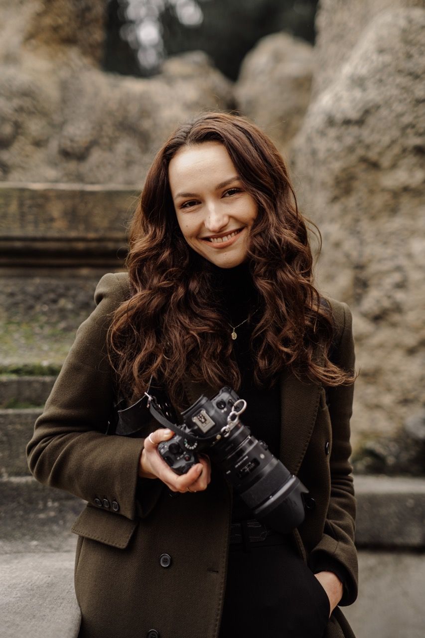 A photographer in a brown coat holding a professional camera stands against a stone wall outdoors.