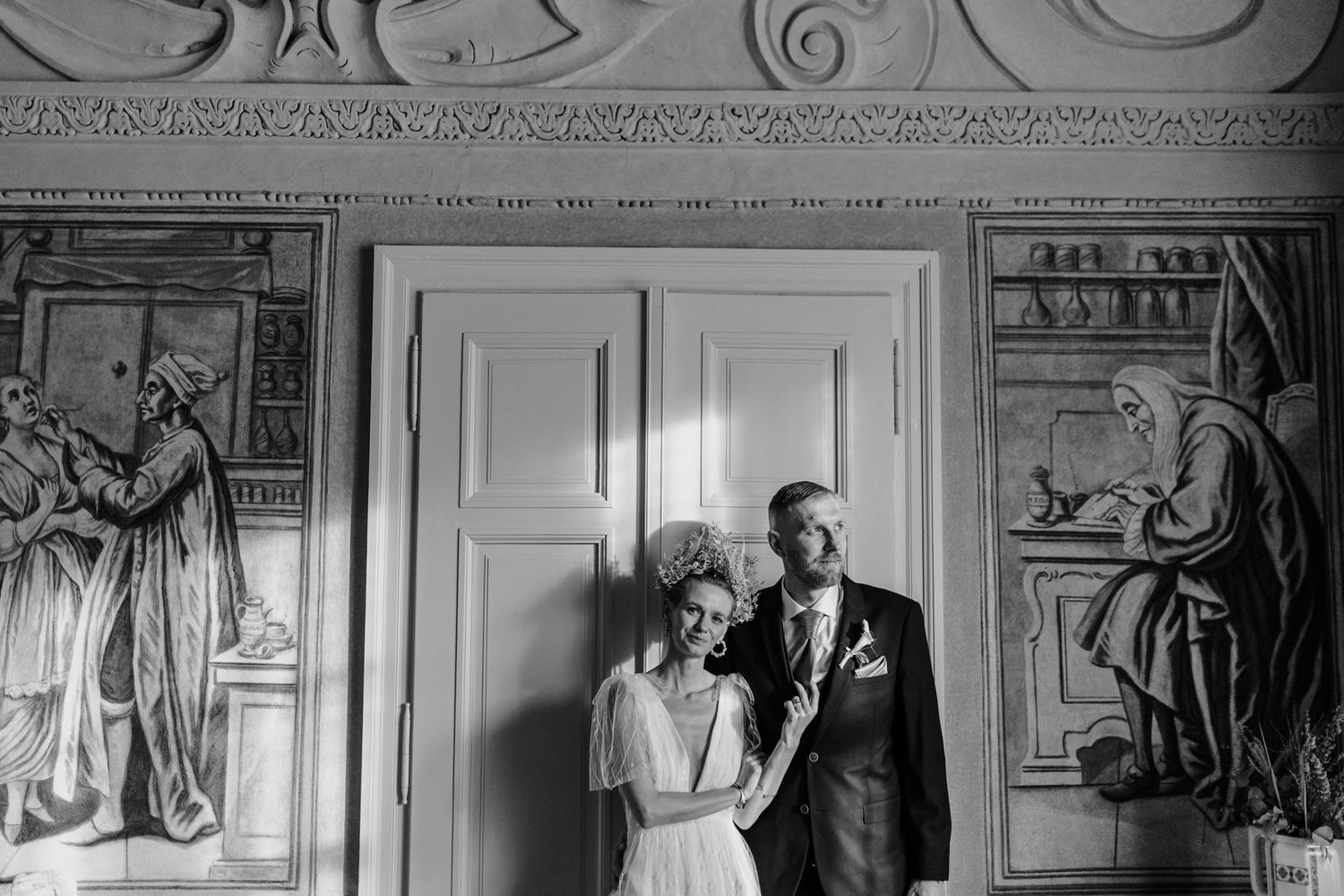 Black and white wedding portrait of couple against wall with classical painted murals.