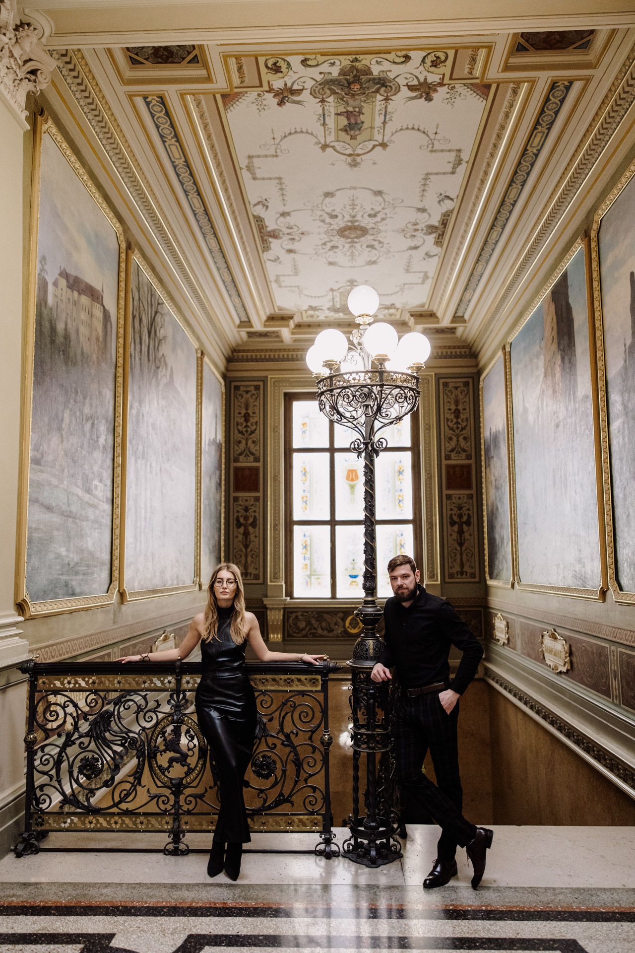 Two people posing in an ornate historic hallway with baroque ceiling, vintage chandelier and decorative iron railing.