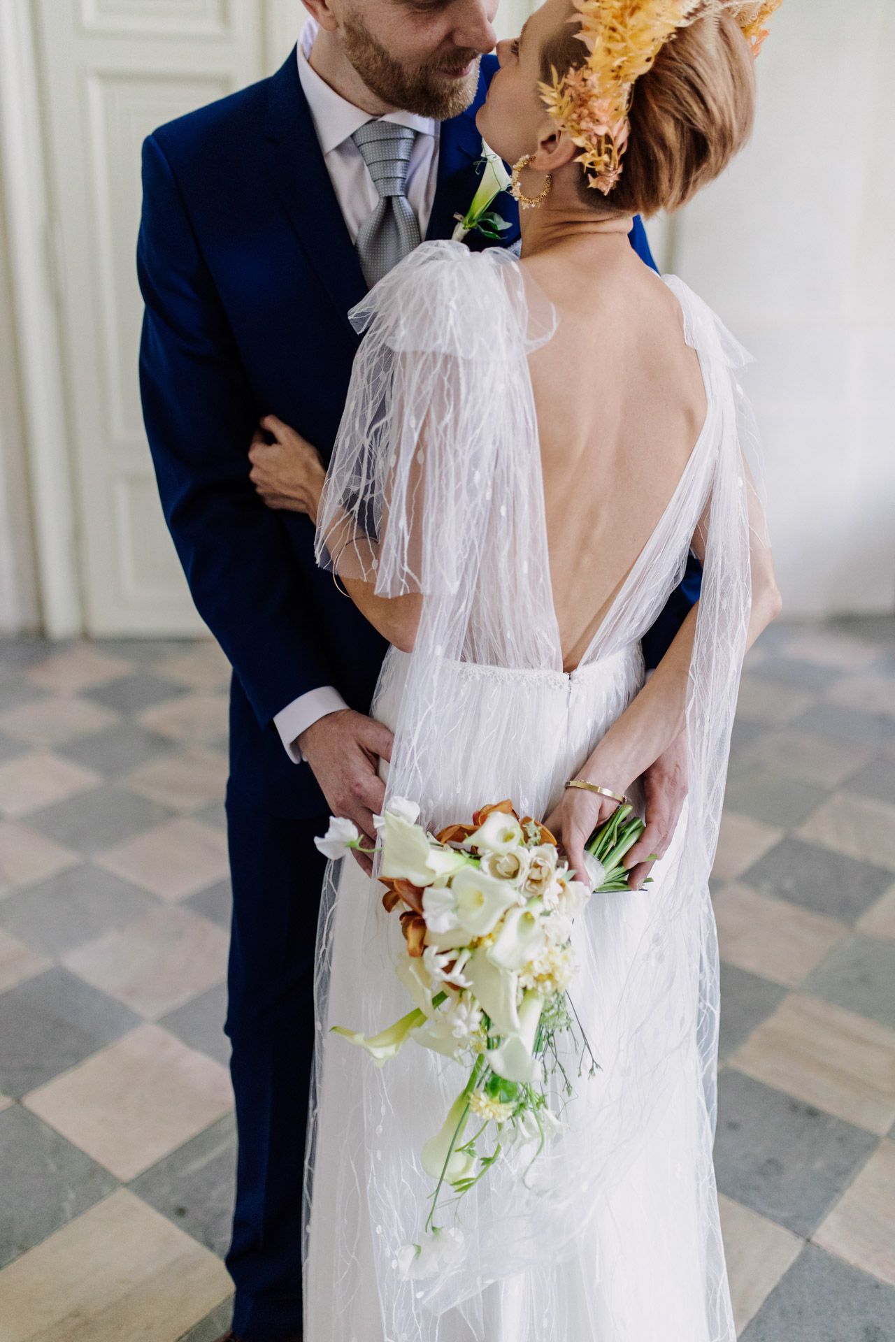 Elegant bride in backless lace gown holding white bouquet while groom in navy suit embraces her.