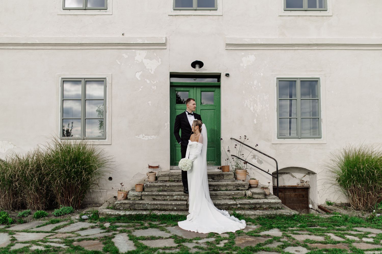 A couple in wedding attire stands on stone steps in front of a white building with a green door and ornamental grasses.