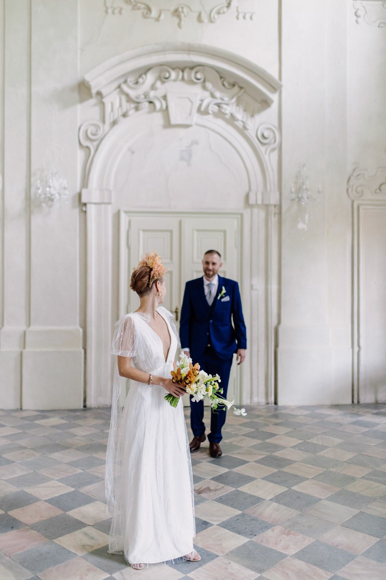 Elegant wedding scene in historic white room with ornate archways and checkered floor.