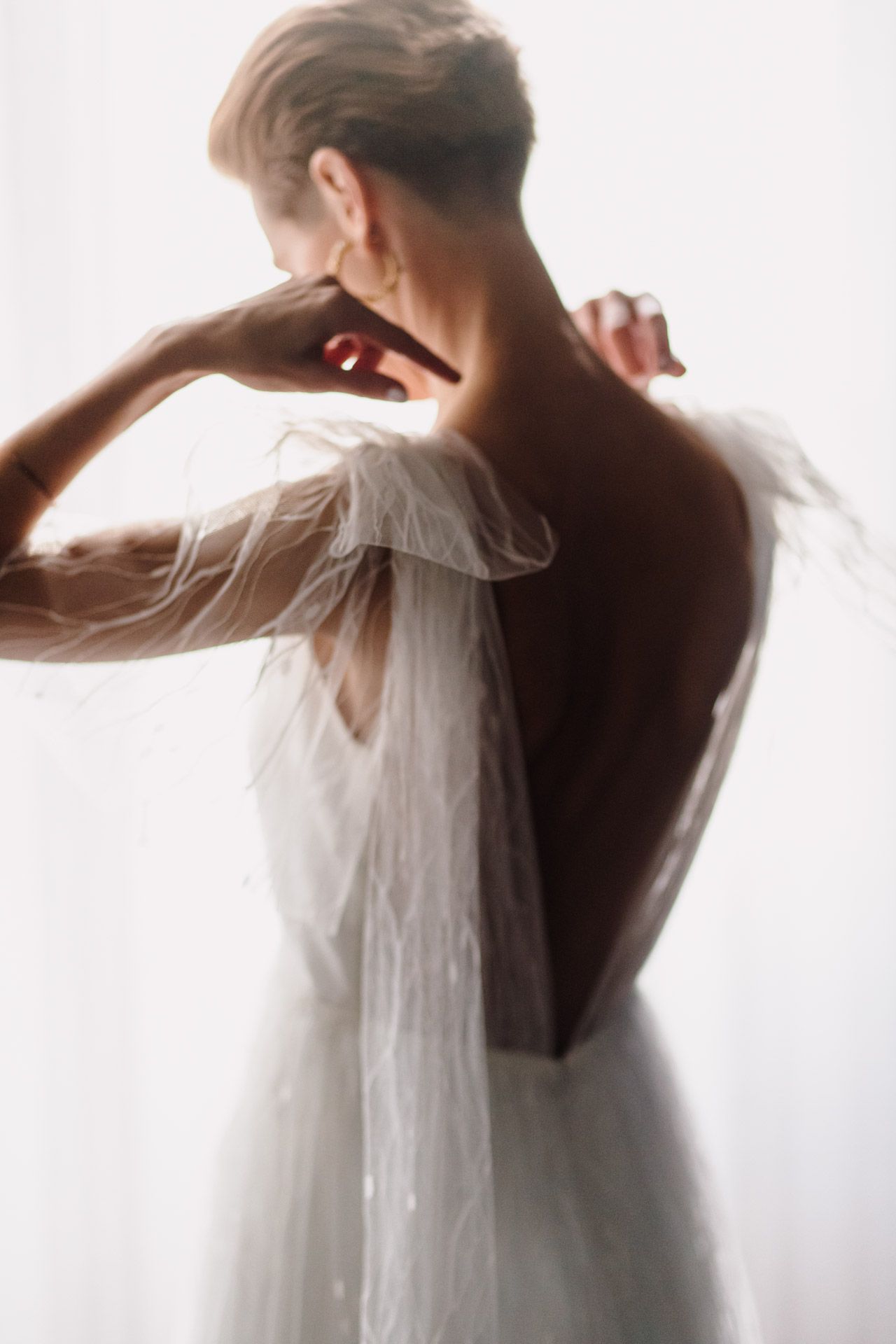 Ethereal back view of bride in delicate white dress with sheer flutter sleeves against bright window.