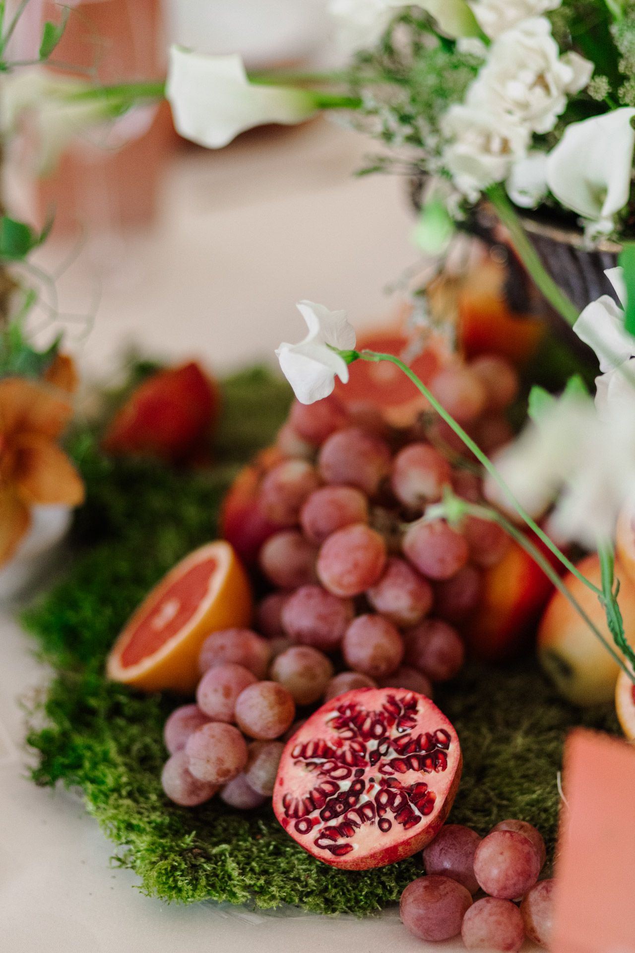 Fresh fruit display featuring purple grapes and a cut pomegranate on moss with delicate white flowers.