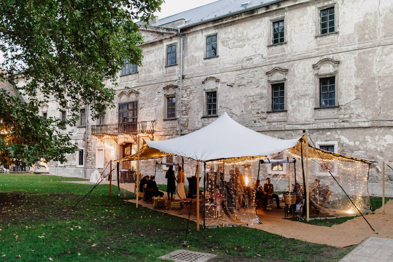 Outdoor wedding reception setup with white tent and string lights against historic stone building.
