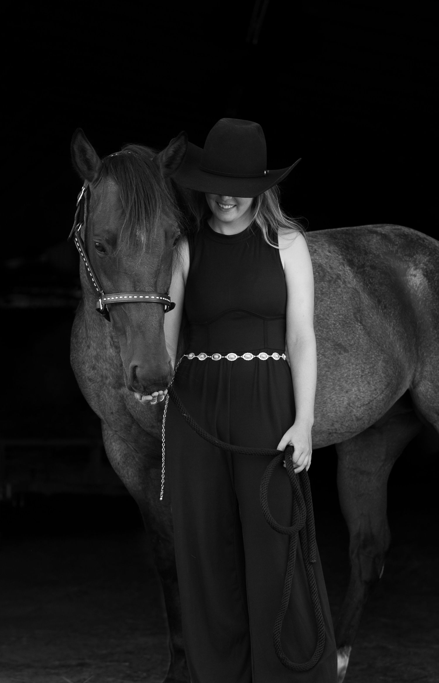 Person in black dress and cowboy hat poses with dark horse in dramatic black and white portrait.
