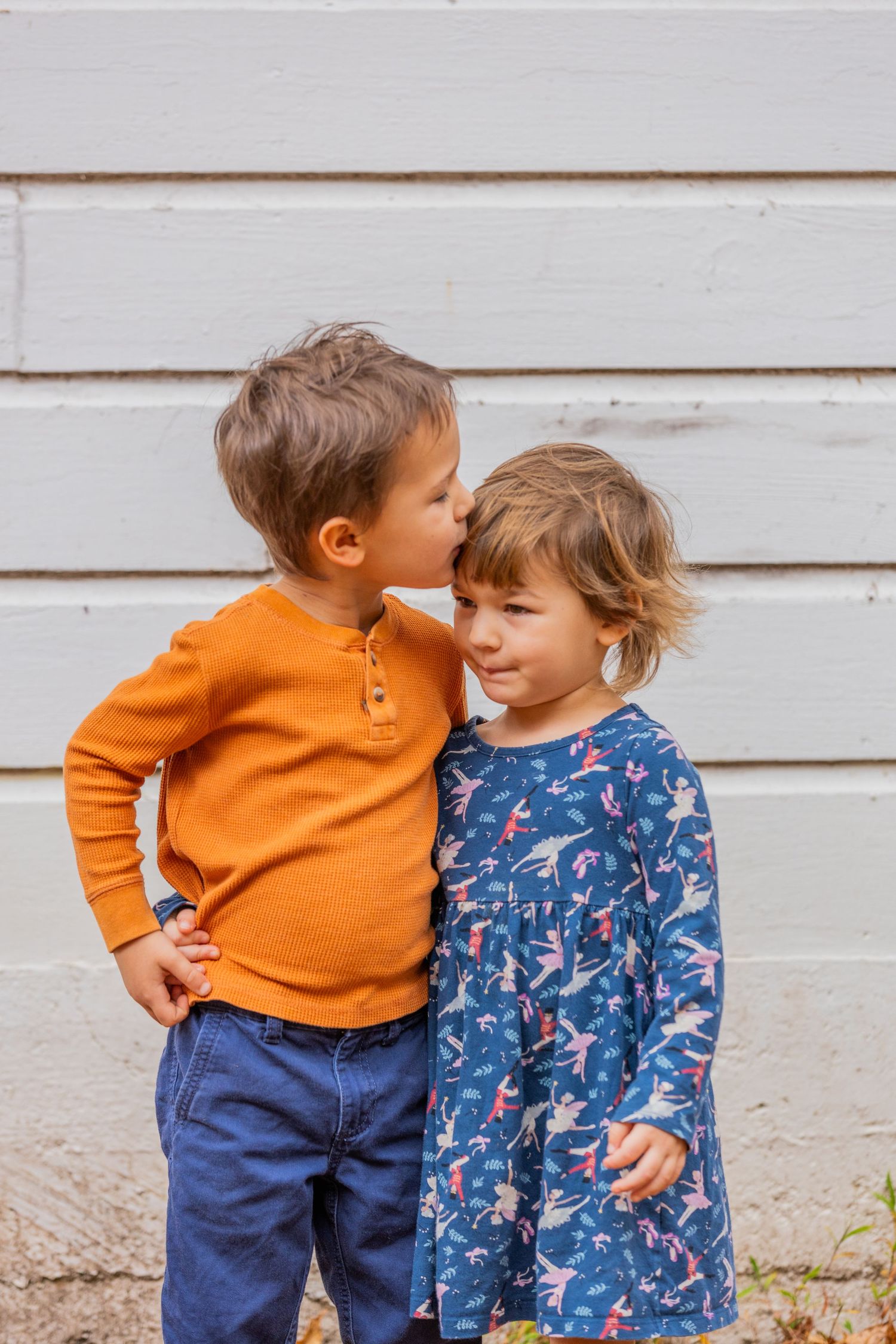 Two young children in colorful sweaters sharing a sweet moment against a white wall.